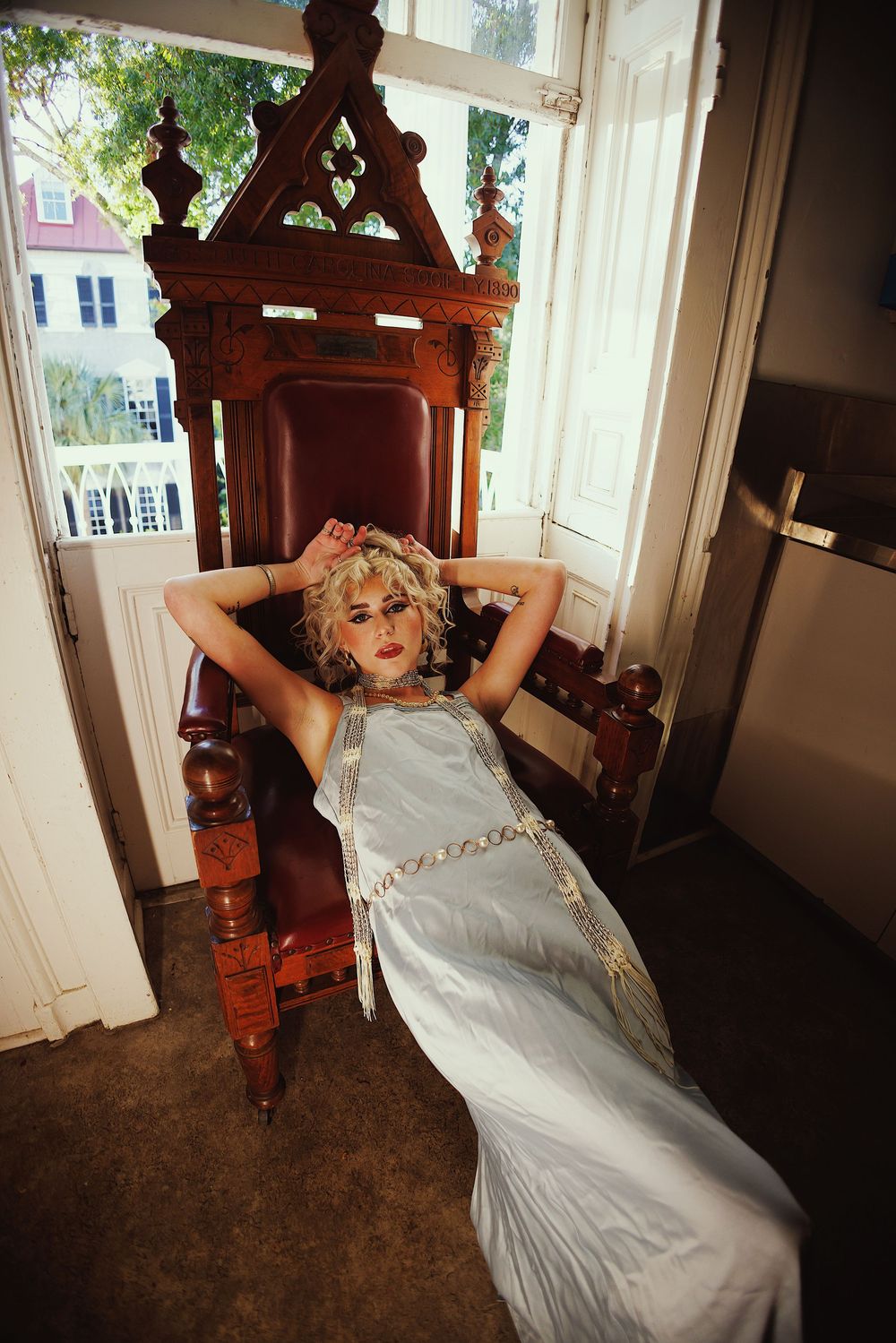 Figure in silver dress lounges dramatically on antique furniture in sunlit room.