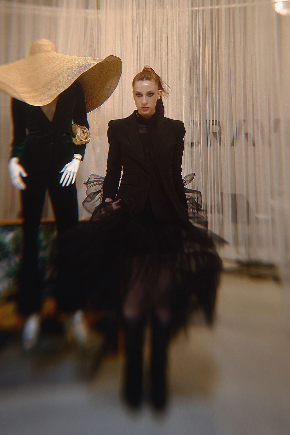 Experimental fashion shoot with blurred figures in black attire against white curtained background.