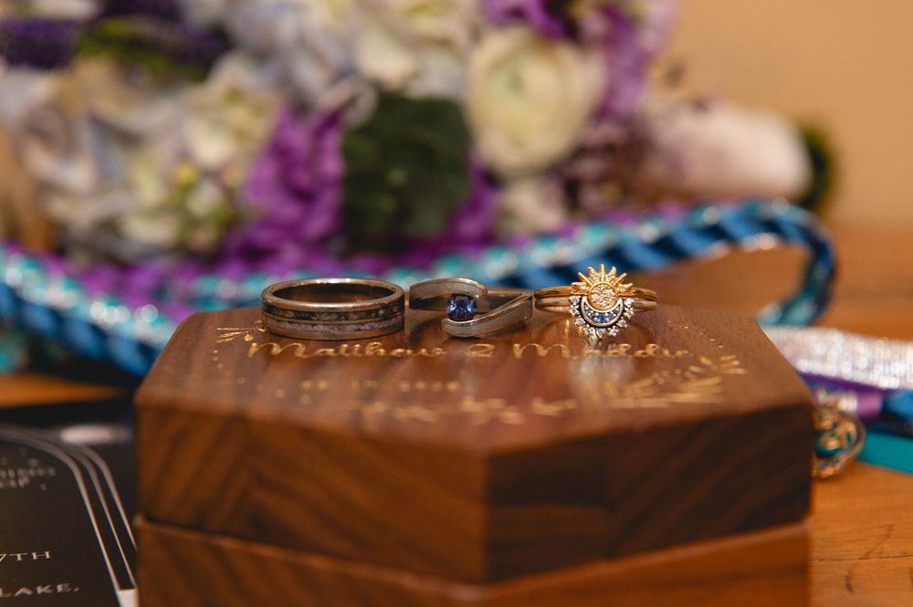 Two wedding rings sit atop a wooden hexagonal box with colorful ribbons and decor in the background.