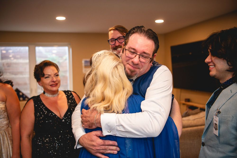 Two wedding guests embrace in an emotional hug while others look on with smiles at an indoor reception venue.