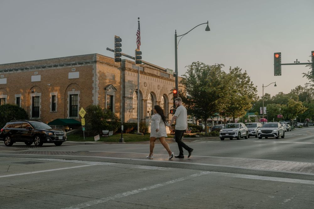 People walk along a city sidewalk with brick buildings and parked cars visible on a cloudy day.
