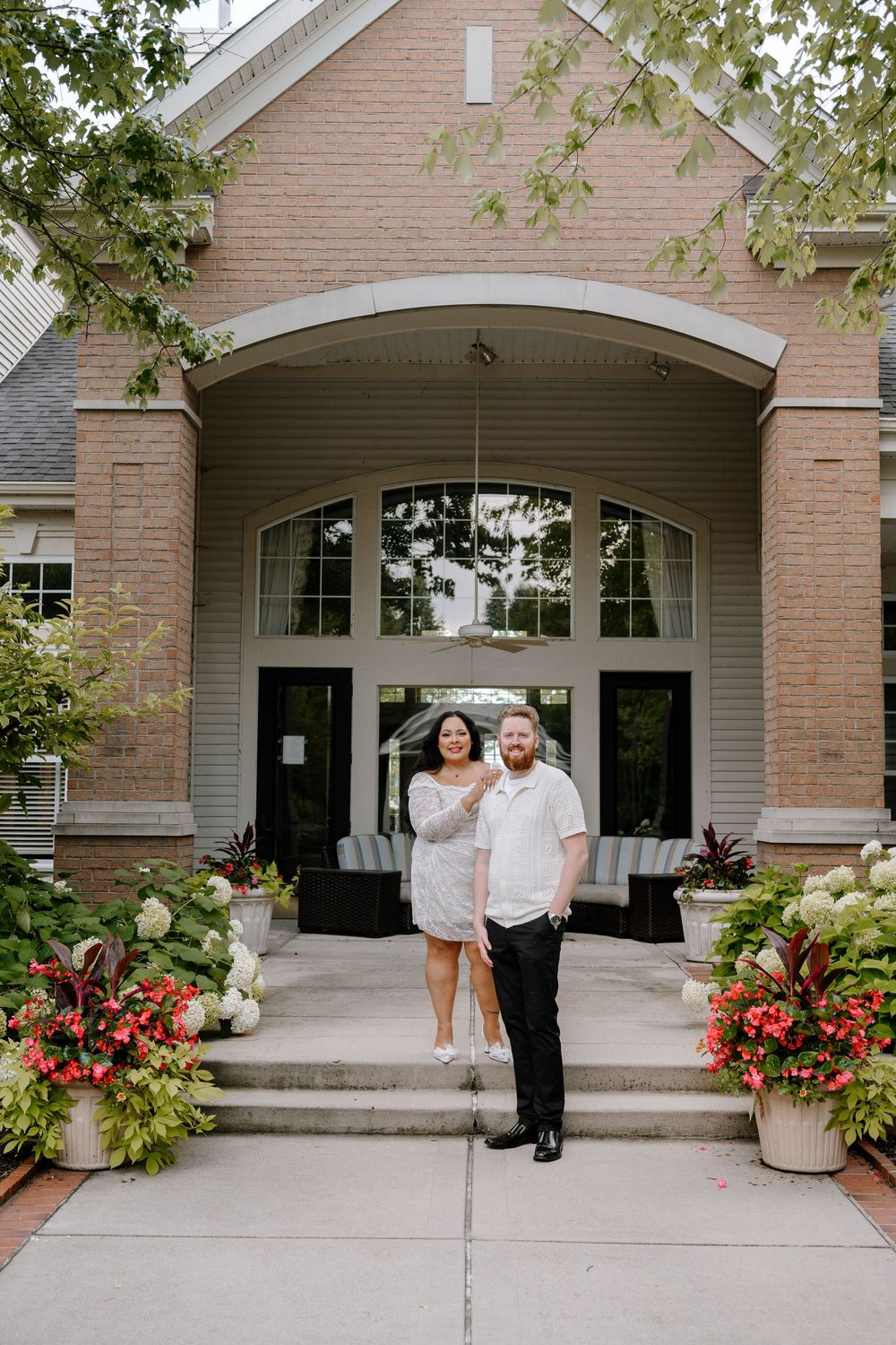 Couple in formal attire standing on steps outside entrance to modern brick building with arched doorway and potted plants.