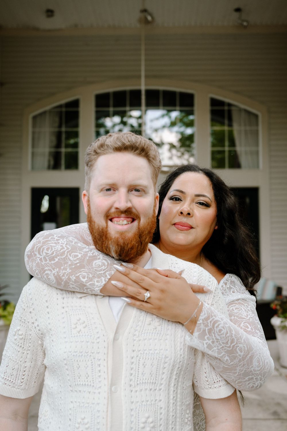 A couple in matching white lace outfits smiles and embraces while standing on a porch.