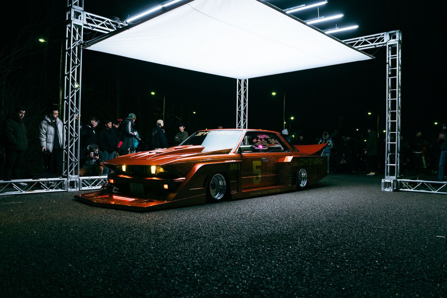 A vintage red Ford Mustang sports car showcased under bright studio lighting on a dark stage with truss rigging.