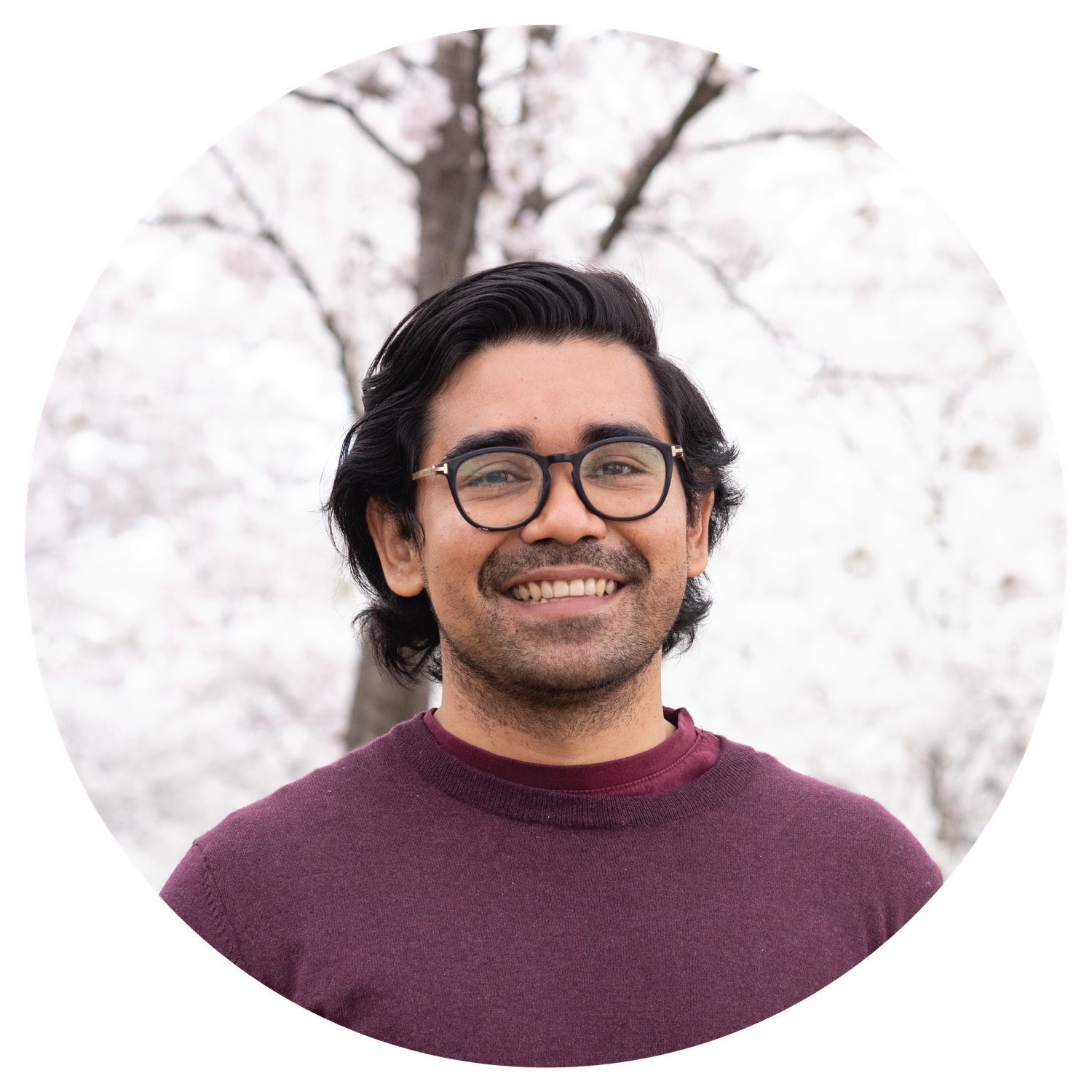 Person wearing glasses and burgundy sweater smiles warmly in front of white cherry blossom trees.