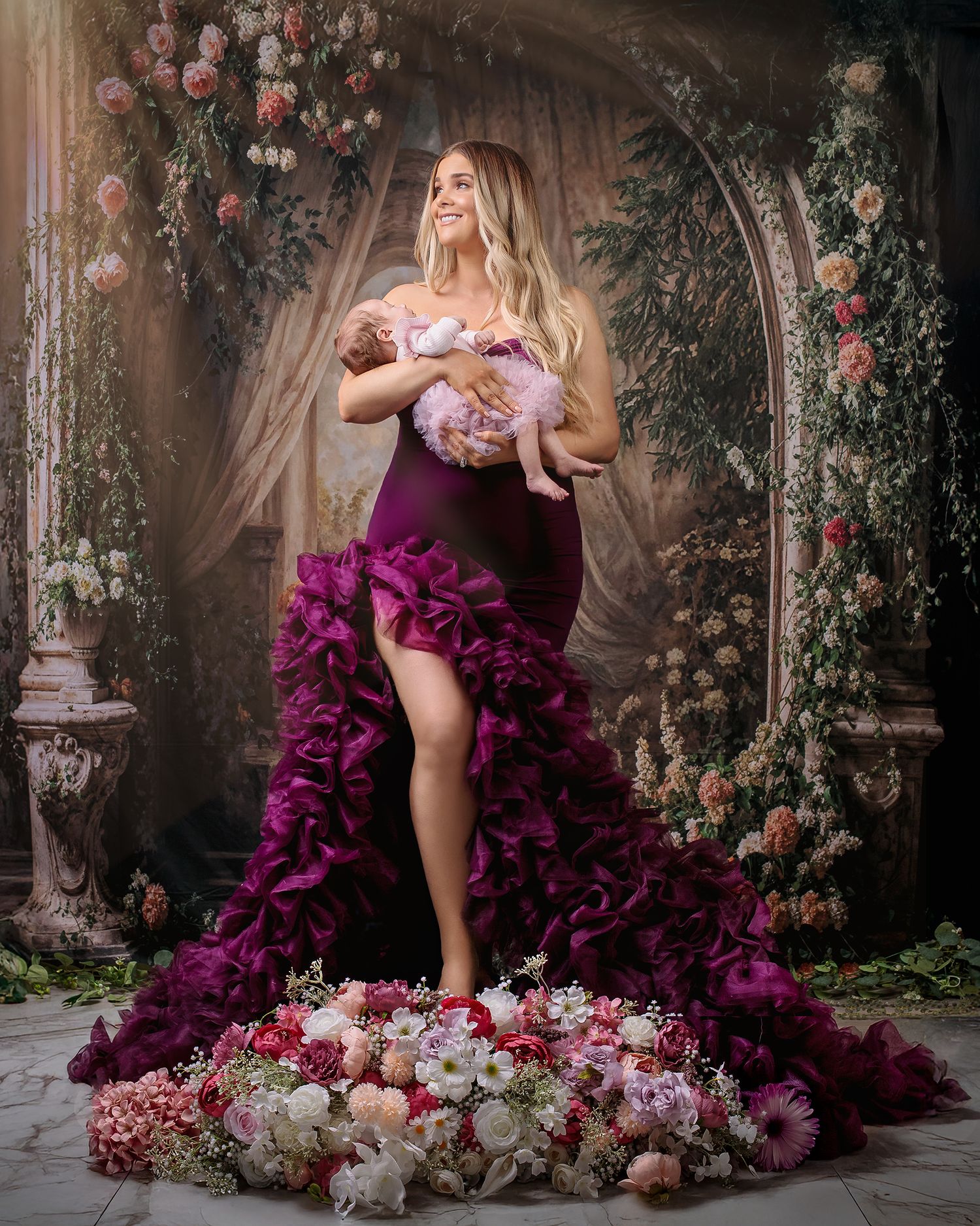 A mother stands on a flower filled podium holding her newborn baby daughter as sunlight beams hit their faces a renaissance arch behind them