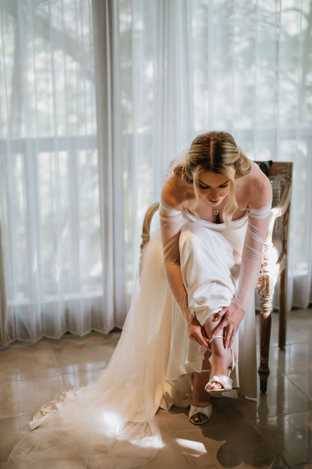 A bride in white adjusts her shoes while sitting in an elegant room with sheer curtains.