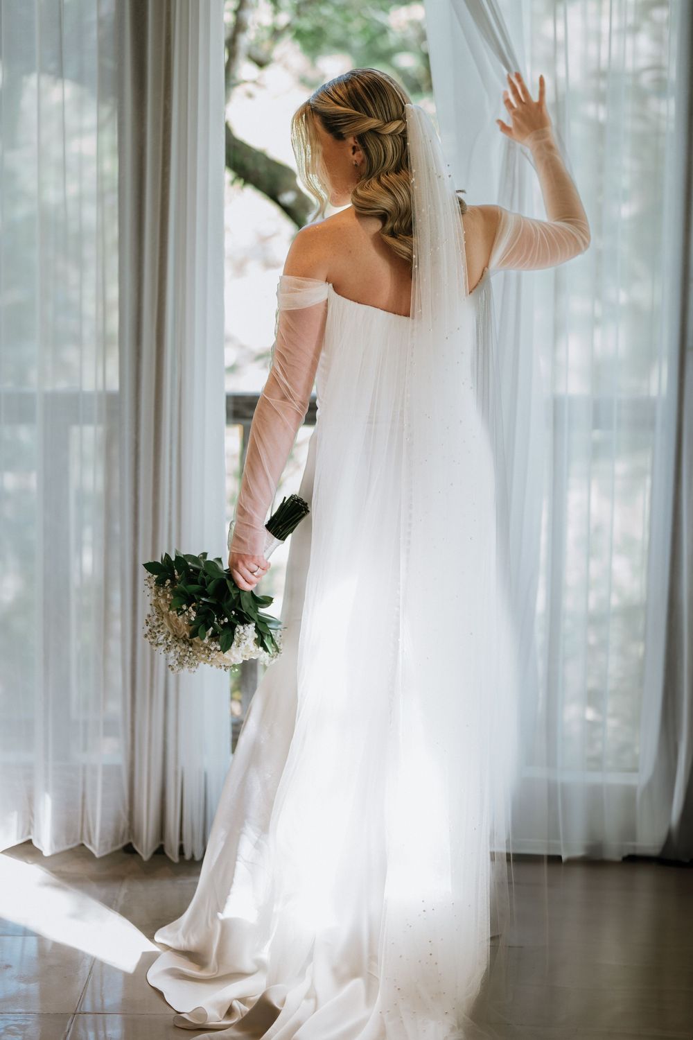 A bride in a flowing white dress twirls in front of sunlit windows.