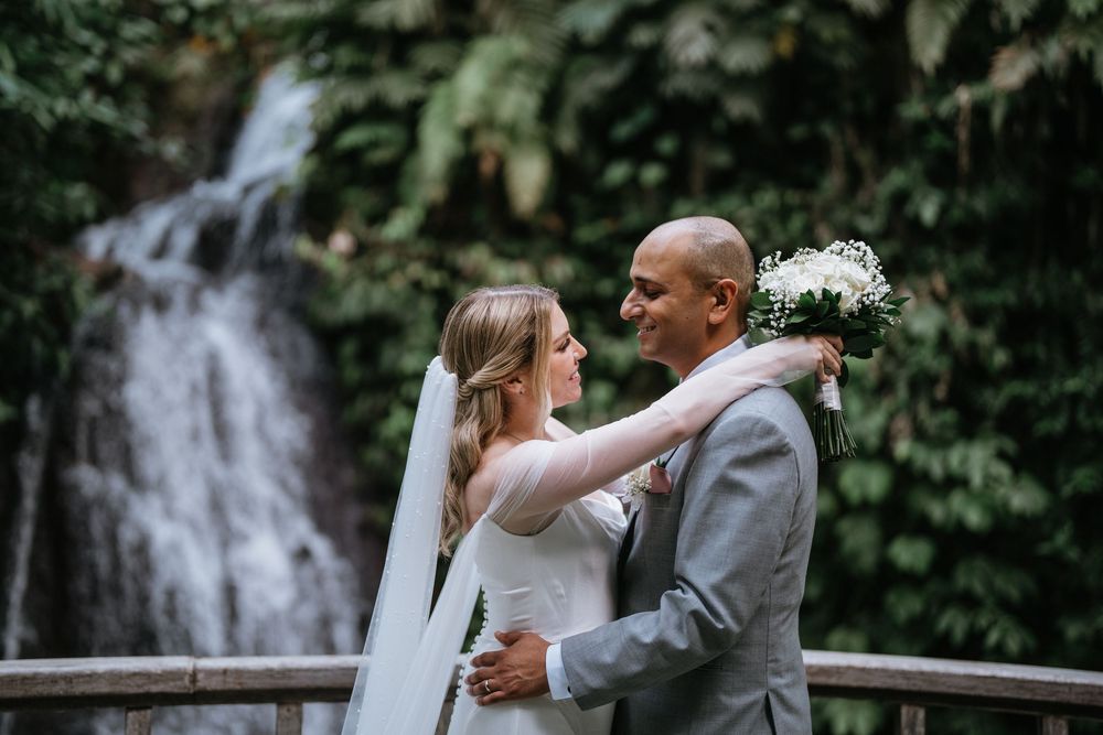 A romantic wedding couple embraces near a cascading waterfall in a lush garden setting.