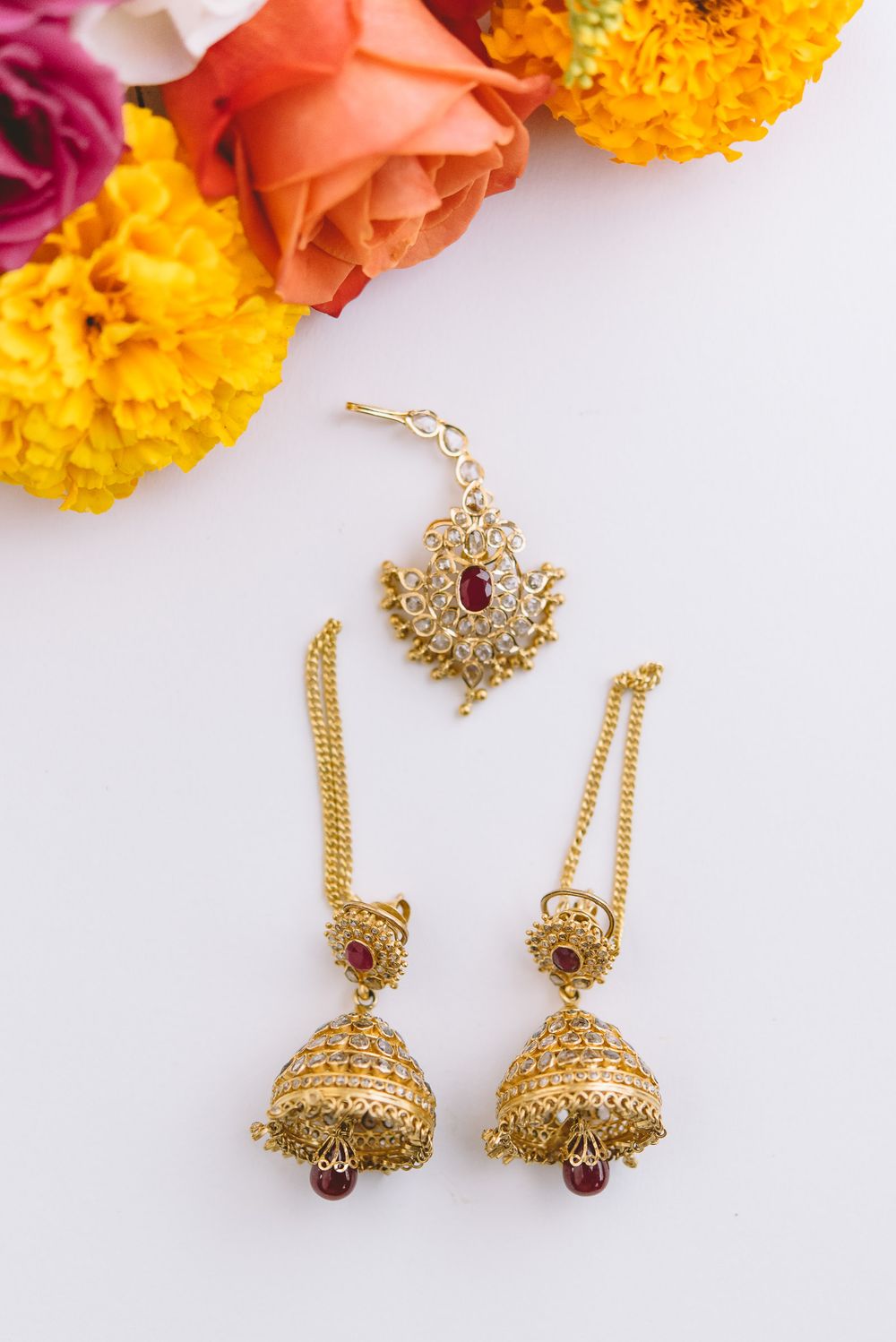 Golden Indian jhumka earrings and tikka set with red stones displayed with pink and yellow marigold flowers.