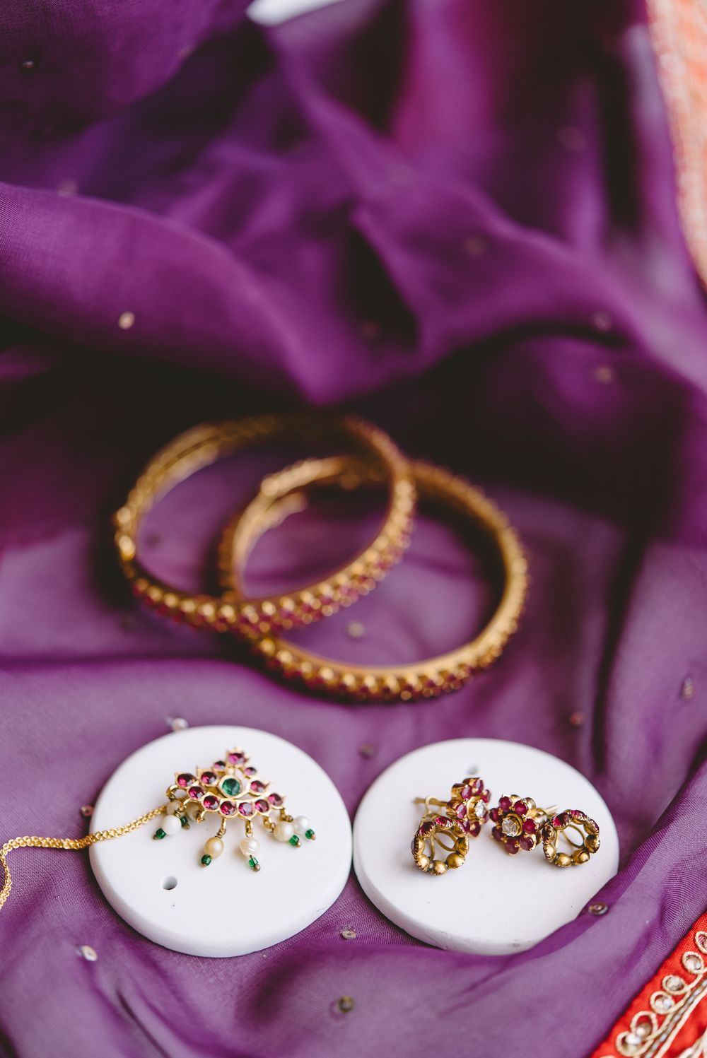 Gold earrings with red gemstones displayed on white circular stands against a rich purple background.
