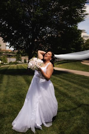 Joyful couple in wedding attire spins and dances together on a green lawn with buildings in the background.