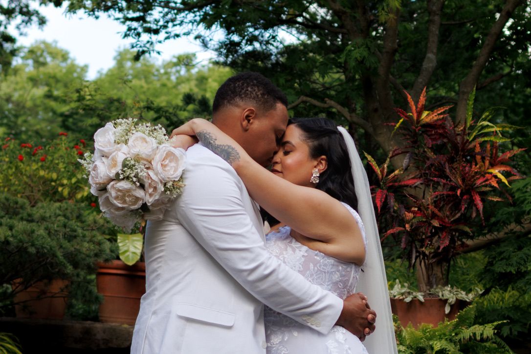 A romantic newlywed couple embraces in a garden setting with lush greenery and a beautiful bridal bouquet of pink roses.