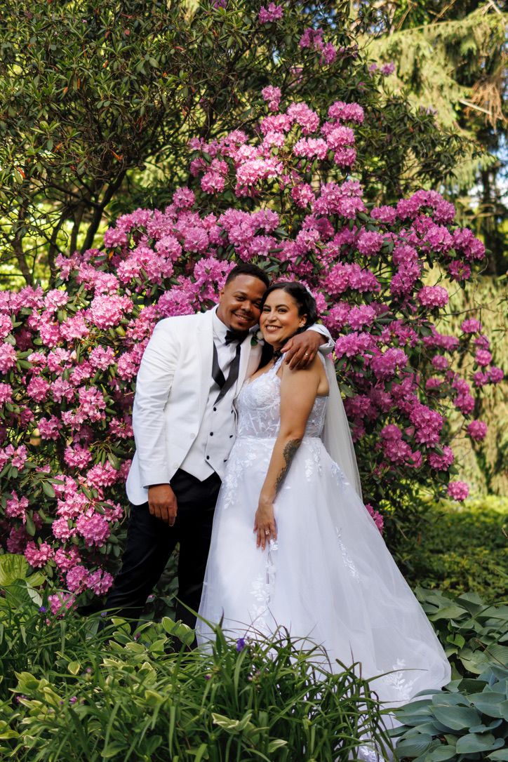 A loving couple in white wedding attire embrace in front of vibrant pink azalea bushes in a garden setting.