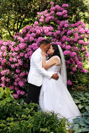 A couple shares a romantic embrace surrounded by vibrant pink azalea bushes in a lush garden setting.