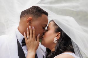 A couple in wedding attire share a romantic kiss under a flowing white veil in an intimate moment.