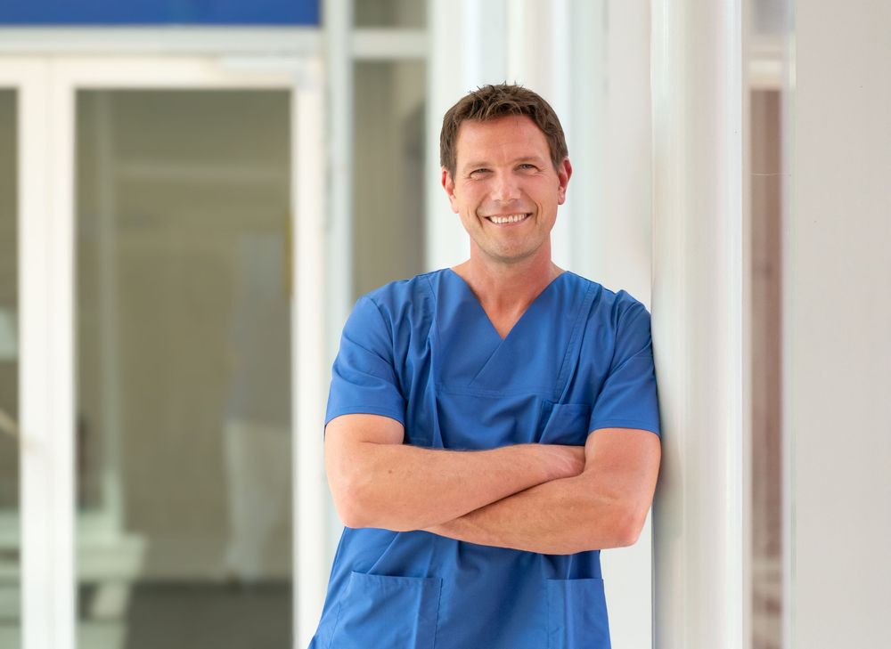 Medical professional in blue scrubs standing in bright hallway with arms crossed.