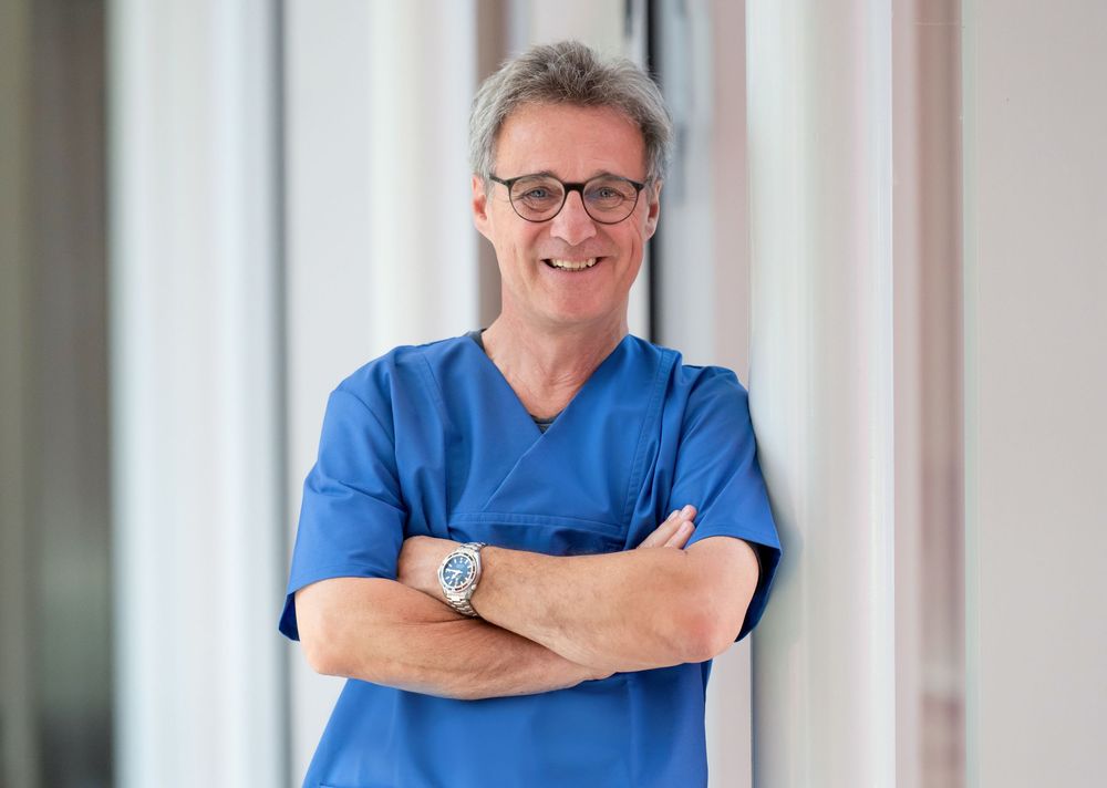 Healthcare worker wearing blue scrubs and glasses posing confidently in medical facility.