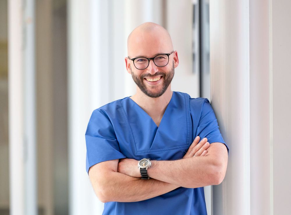 Medical staff member in blue uniform standing with crossed arms in hospital corridor.