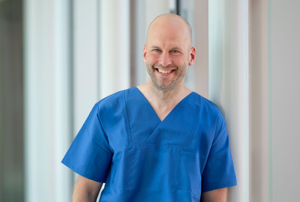 Hospital staff member in blue uniform standing confidently in bright corridor.
