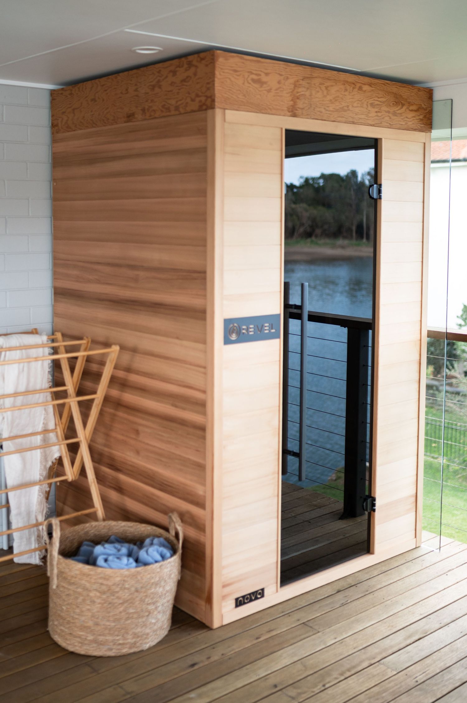 Modern cedar sauna installation on wooden deck with towel rack and woven basket, featuring glass door and lakefront view.