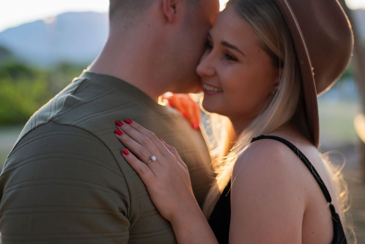 A romantic couple embraces during golden hour, with a woman in a brown hat and black top touching her partner's shoulder.