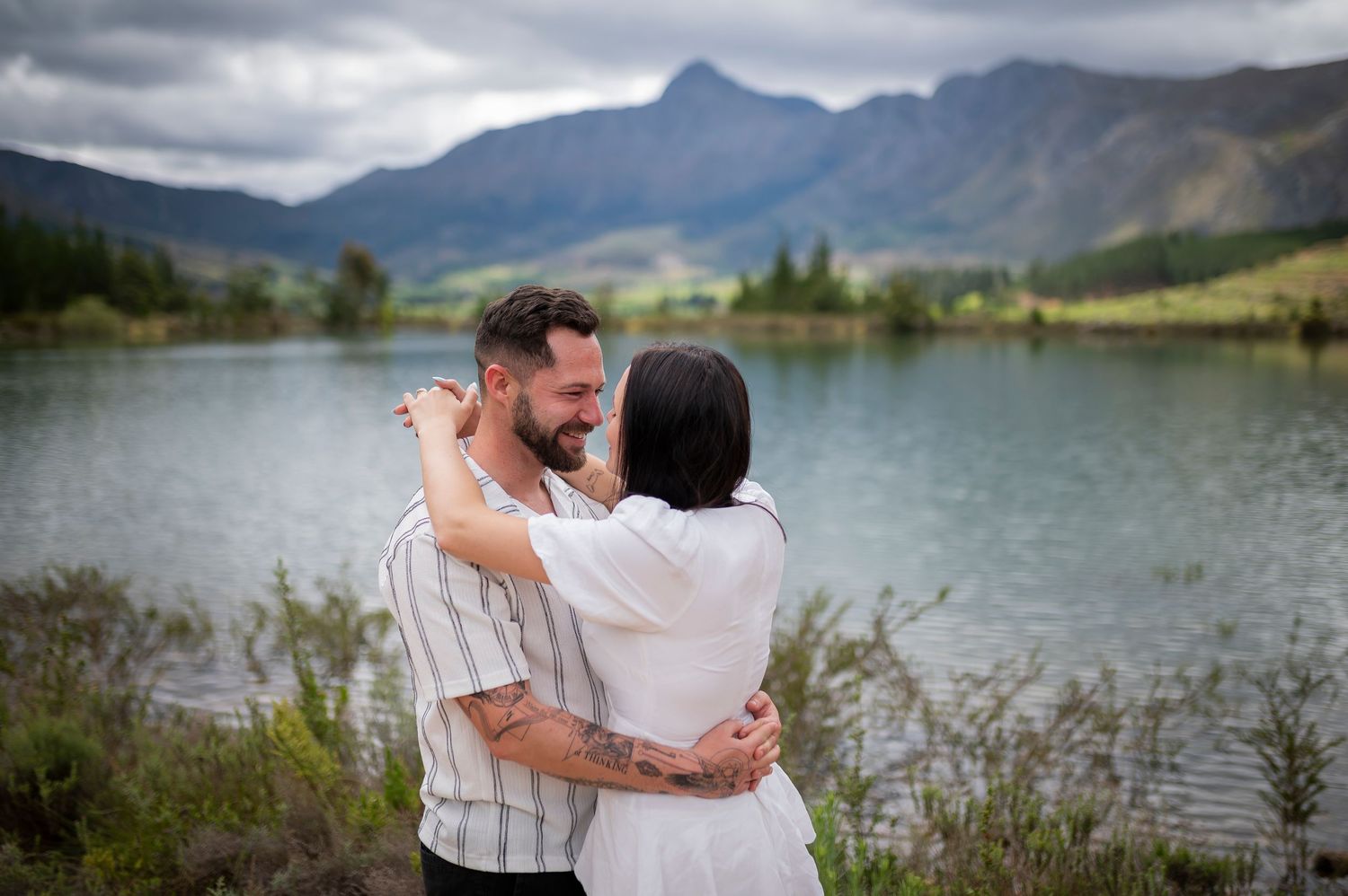 A romantic couple embraces near a scenic mountain lake with cloudy skies in the background.