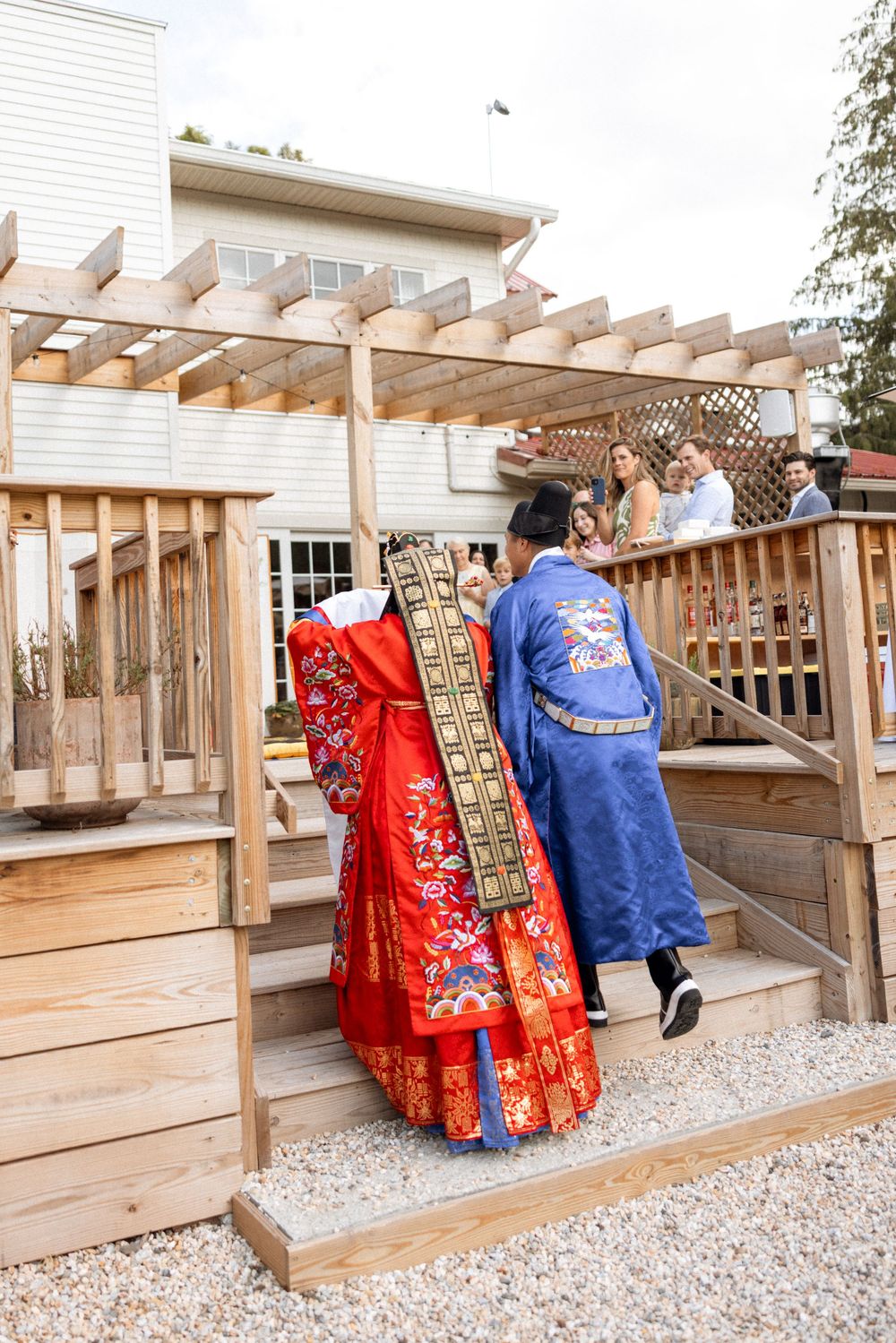 Couple in traditional Korean wedding attire walking along wooden deck with pergola.