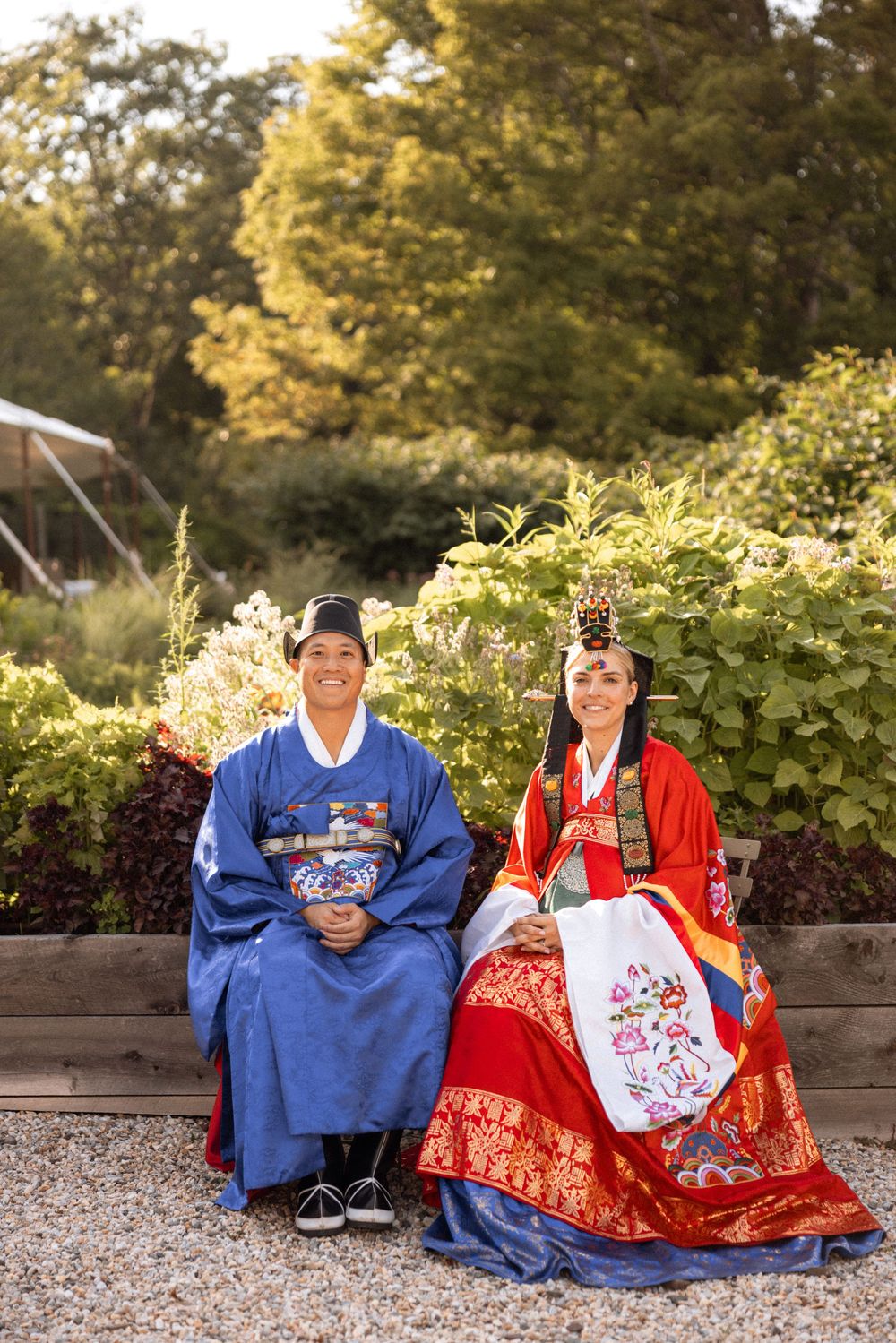 Couple in traditional Korean wedding attire seated together in garden setting.