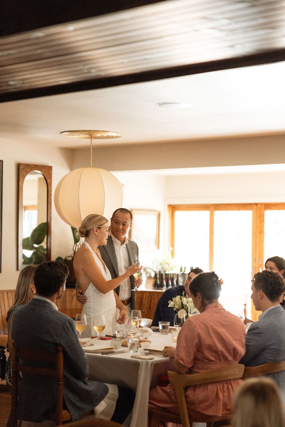 Wedding guests gathered around a dining table with a paper lantern pendant light overhead during an intimate reception.