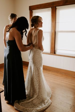 A bride in a lace gown gets help with her dress in a sunlit room with wooden floors.