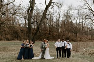 Winter wedding party poses outdoors among bare trees in natural setting.