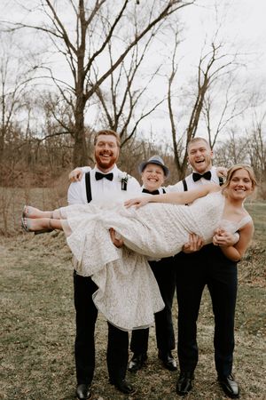 Groomsmen in black suits playfully lift and hold someone in a white dress at an outdoor wedding photoshoot.