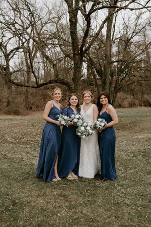 Bridesmaids in navy blue dresses pose with bride in white lace gown holding blue bouquets outdoors.