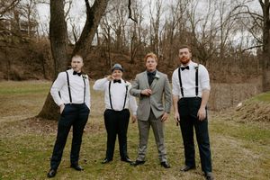 Groomsmen in suspenders and suits pose together on a grassy hillside.