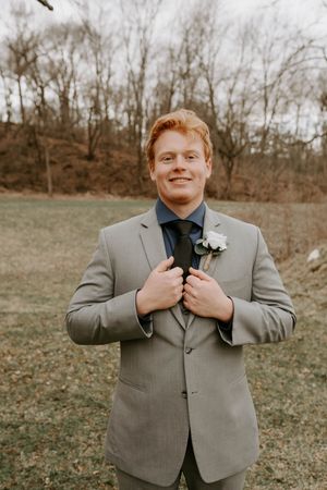 Person in grey suit adjusts tie and poses for wedding photos outdoors.