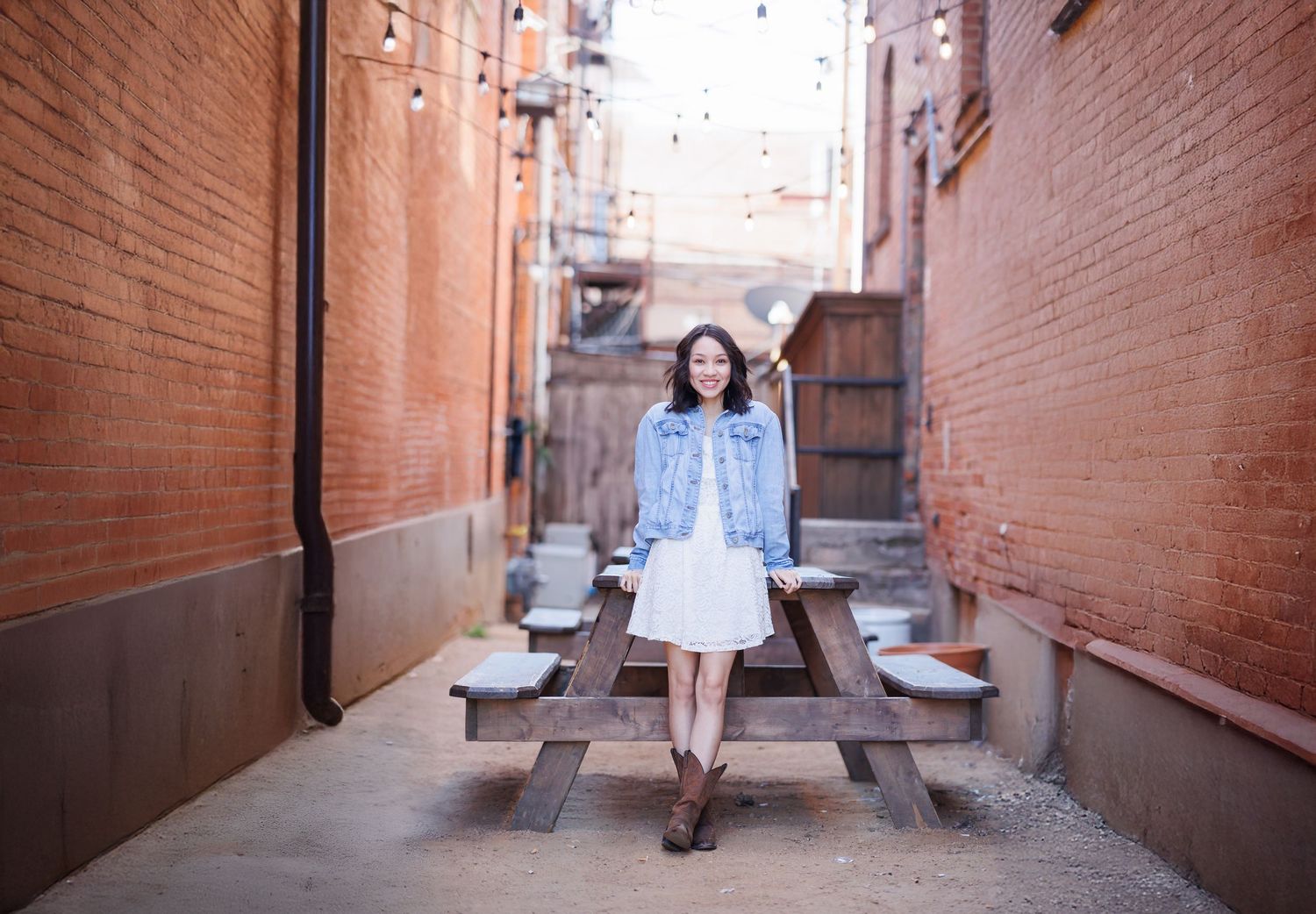 High School Senior sits on a wooden bench between brick buildings in an urban alley in Fort Worth Stockyards for graduation photos