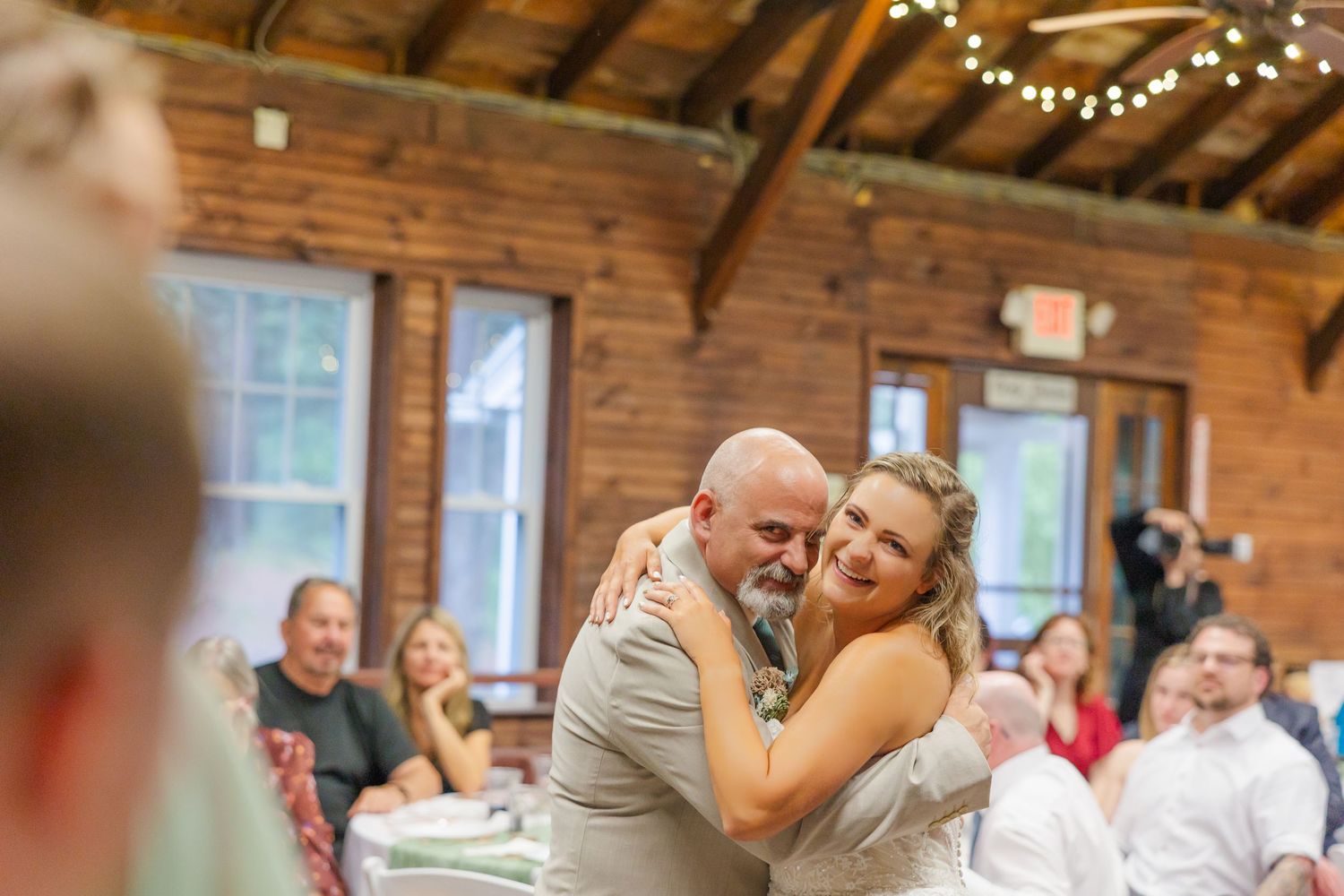 A couple shares an emotional dance moment at a rustic indoor reception venue decorated with string lights.