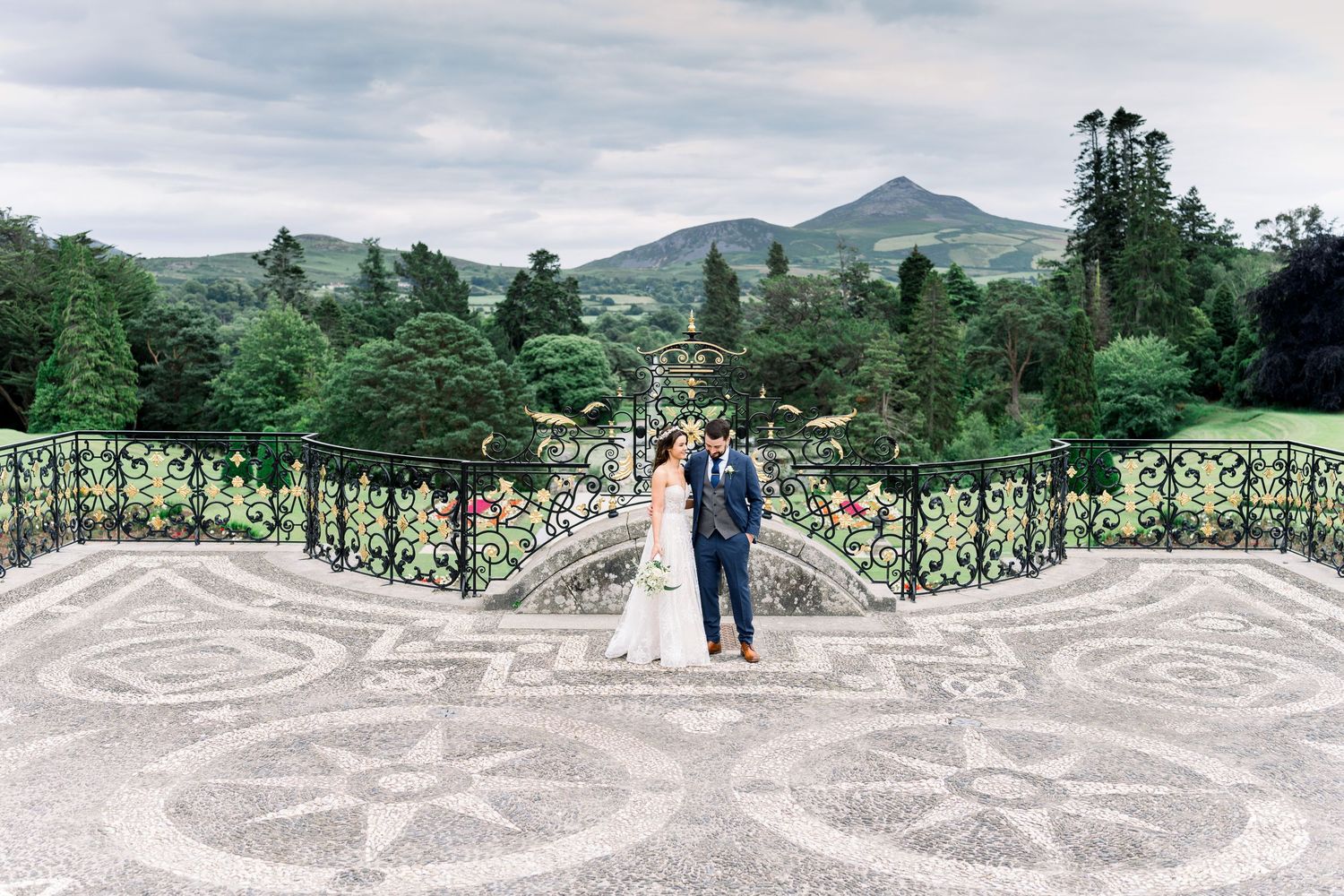A wedding couple stands on an ornate patterned terrace with lush mountains and trees in the background.