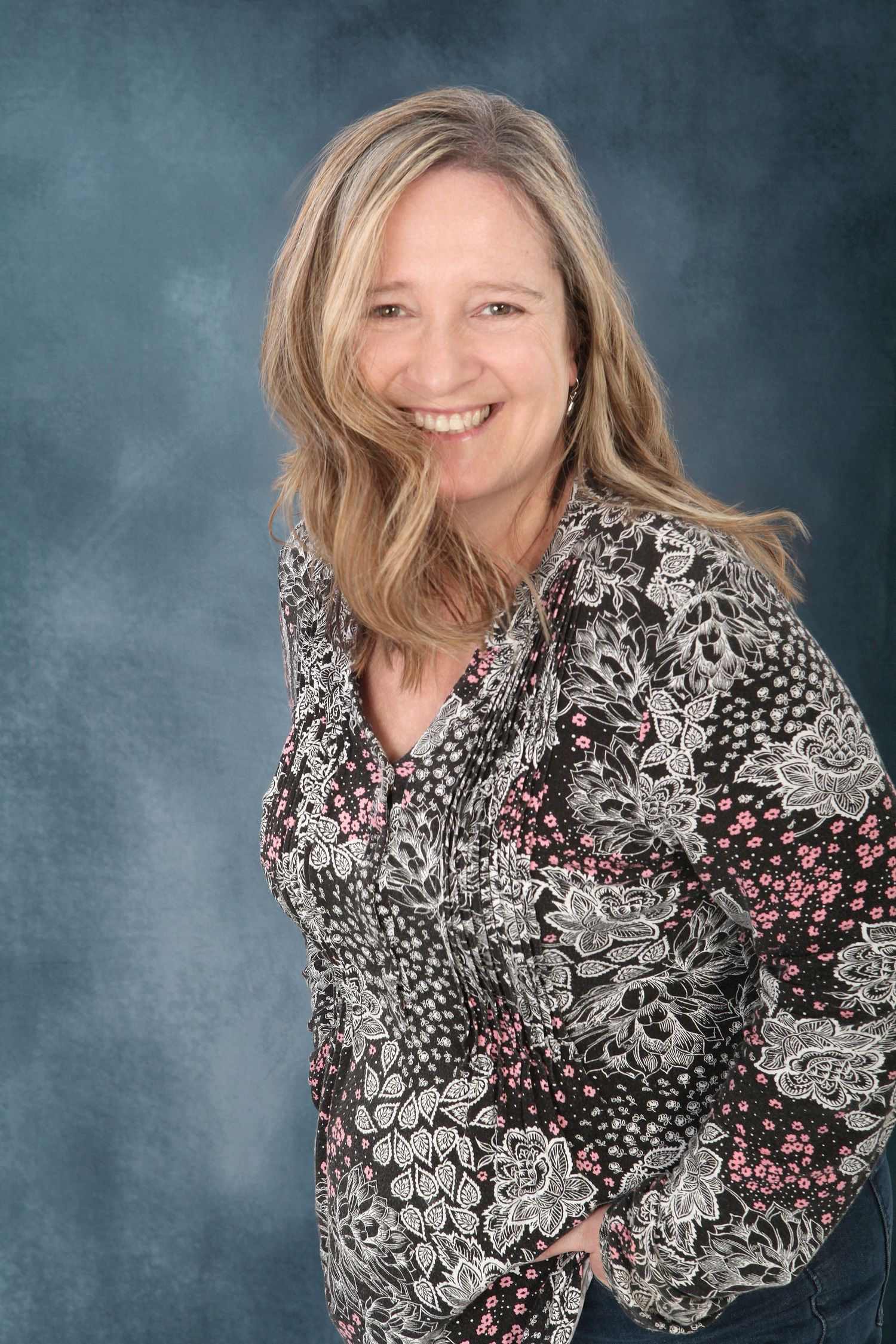 Professional studio portrait of a person in a black and gray floral print blouse smiling against a mottled blue background.