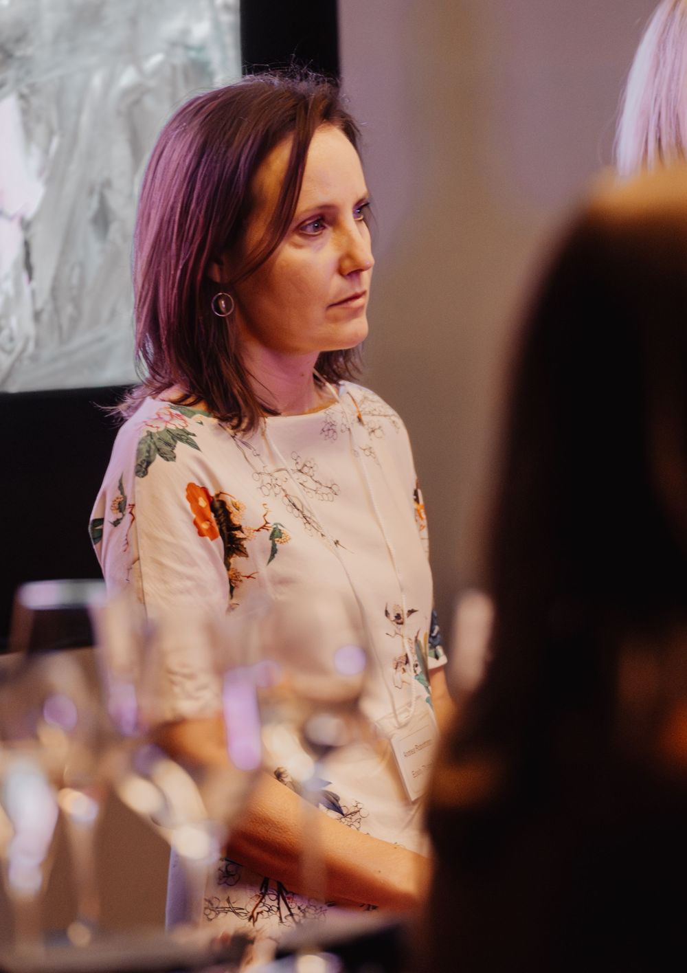 A person with burgundy hair wearing a white jacket sits at a table during what appears to be a formal dinner event.