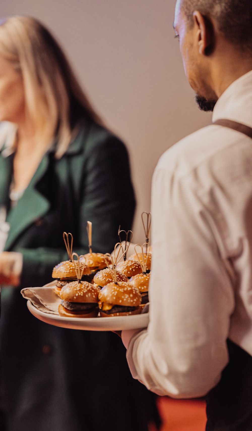 Server in white shirt holding plate of gourmet mini burgers with toothpicks at upscale catering event.