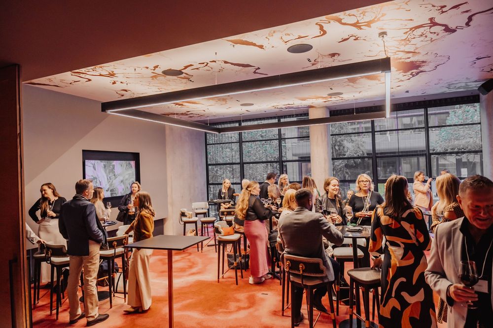 Large open-plan office space with multiple people working at desks and gathering in meeting areas under modern lighting.