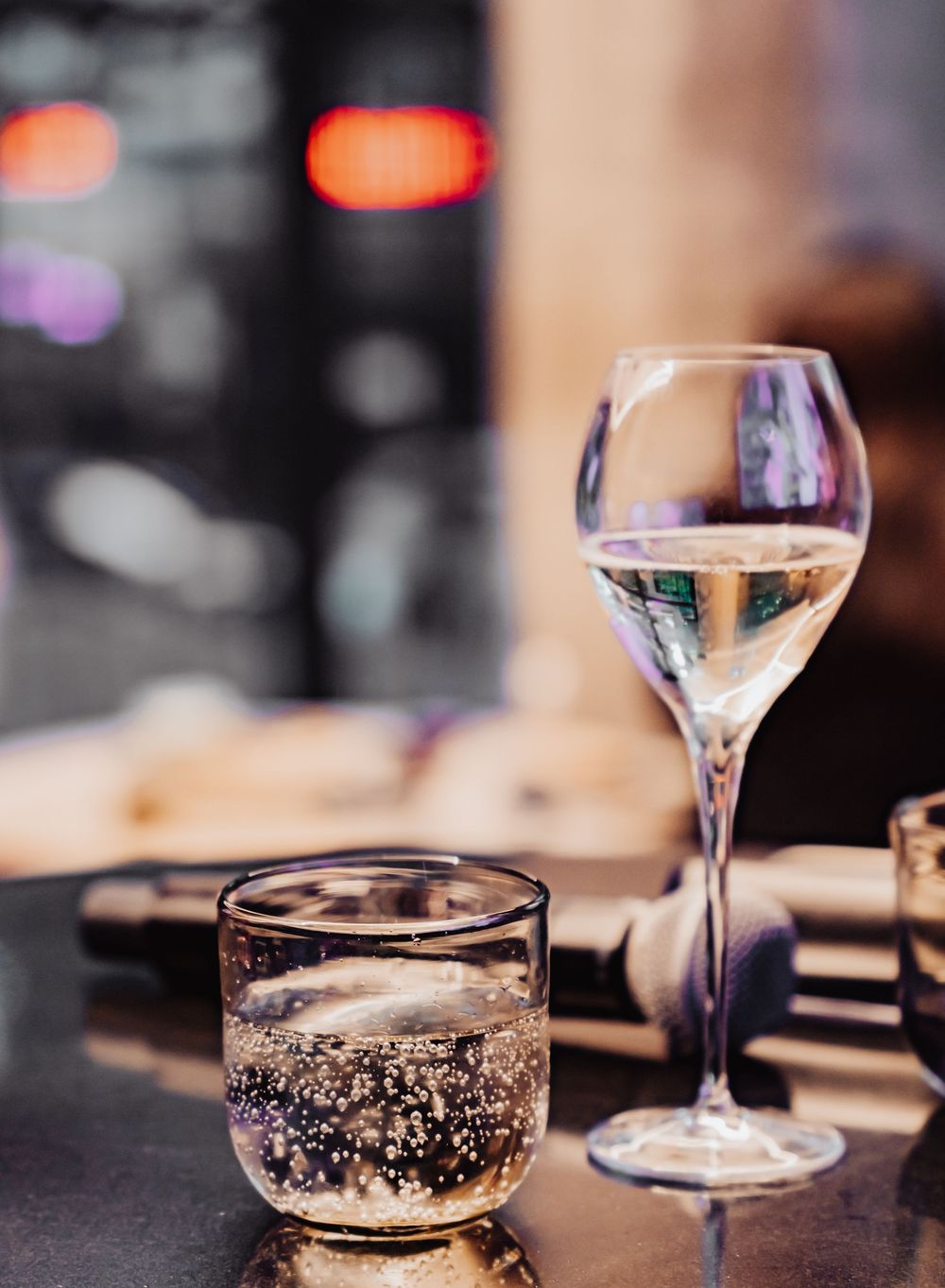 Two elegant glasses with clear sparkling beverages on a dark table with bokeh lights in the background.