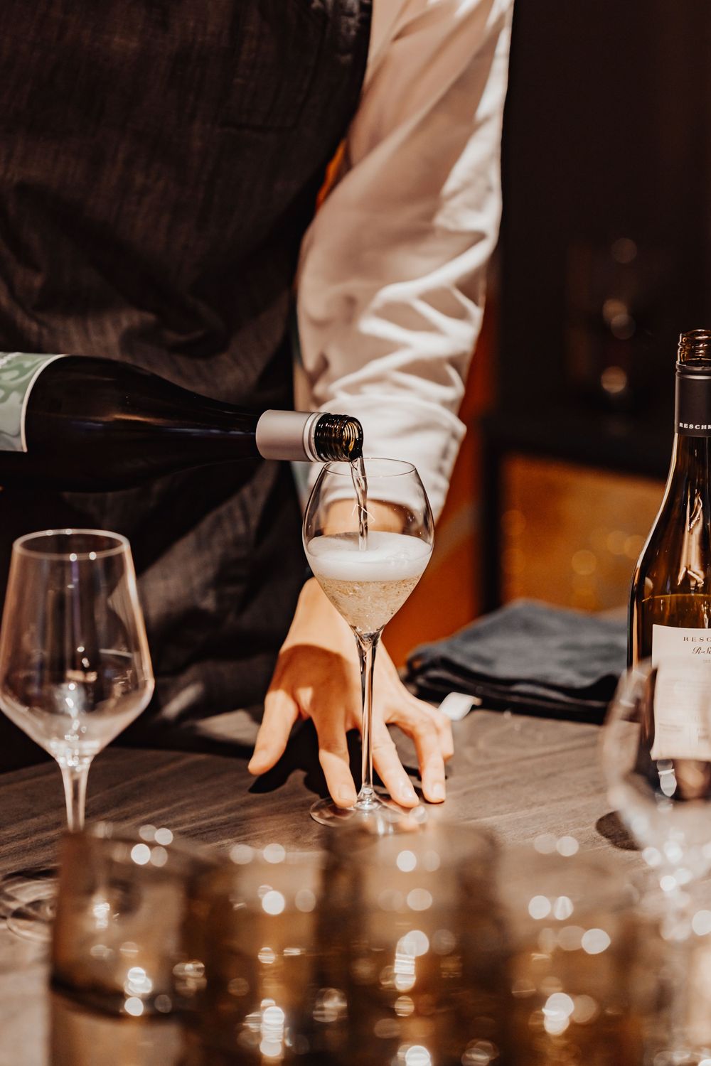 Wine being poured into a glass by someone wearing a white sleeve against a dark background with elegant glassware.