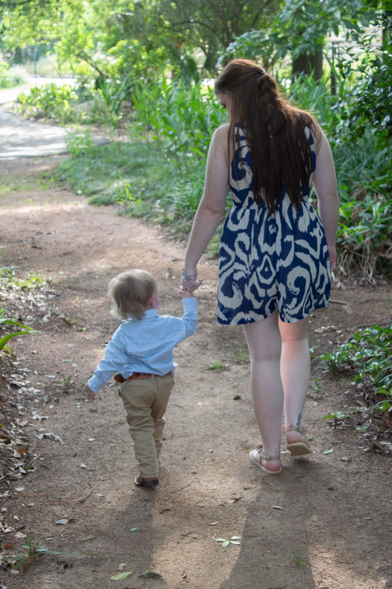 A child and adult walk together along a winding dirt path through green foliage on a sunny day.