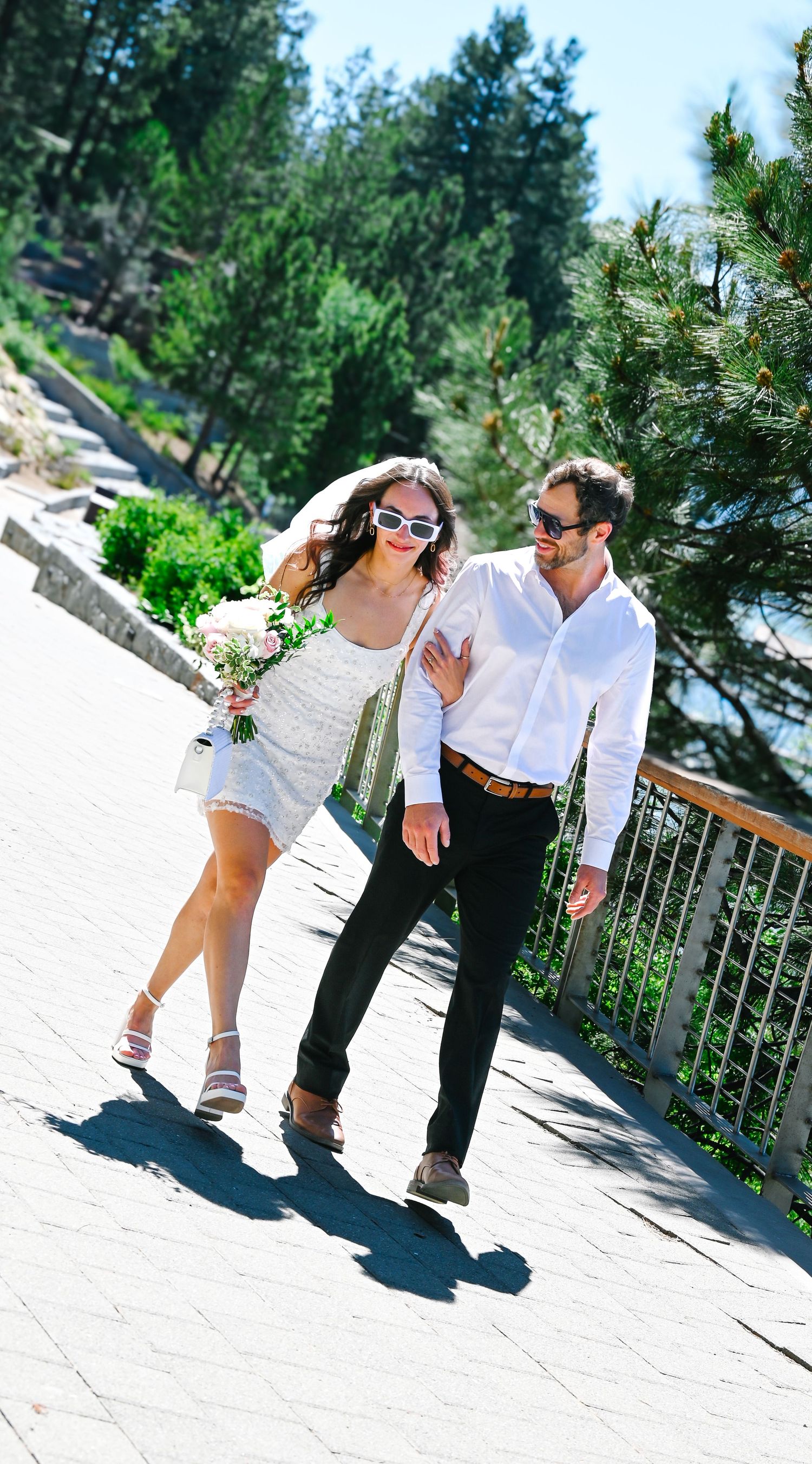 A couple in white outfits playfully dancing and laughing together on a sunny outdoor terrace with trees in the background.