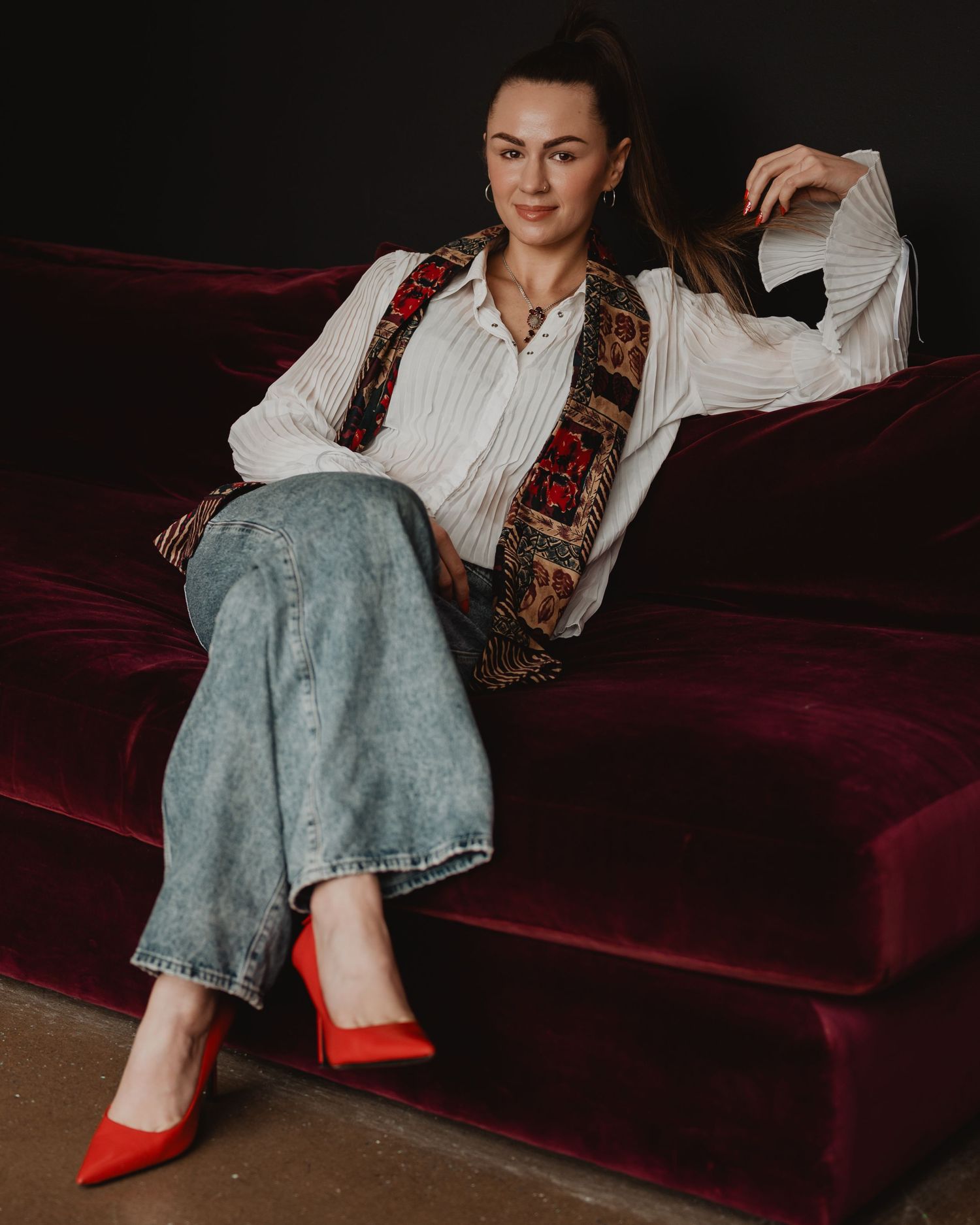 Retro-styled fashion portrait on burgundy velvet couch featuring white shirt, floral vest, jeans and red heels.
