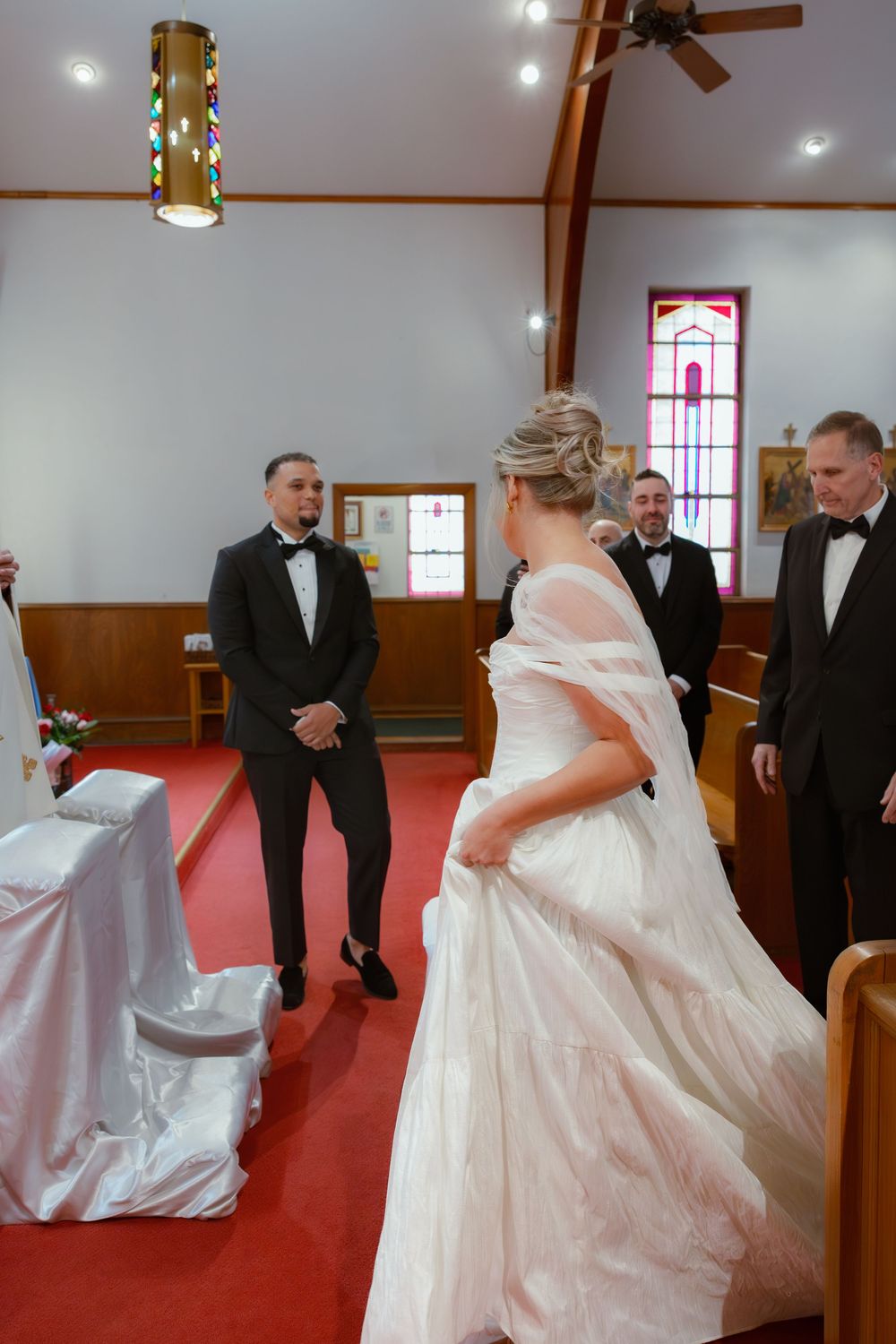 A wedding ceremony takes place inside a small chapel with red carpeting and stained glass windows.