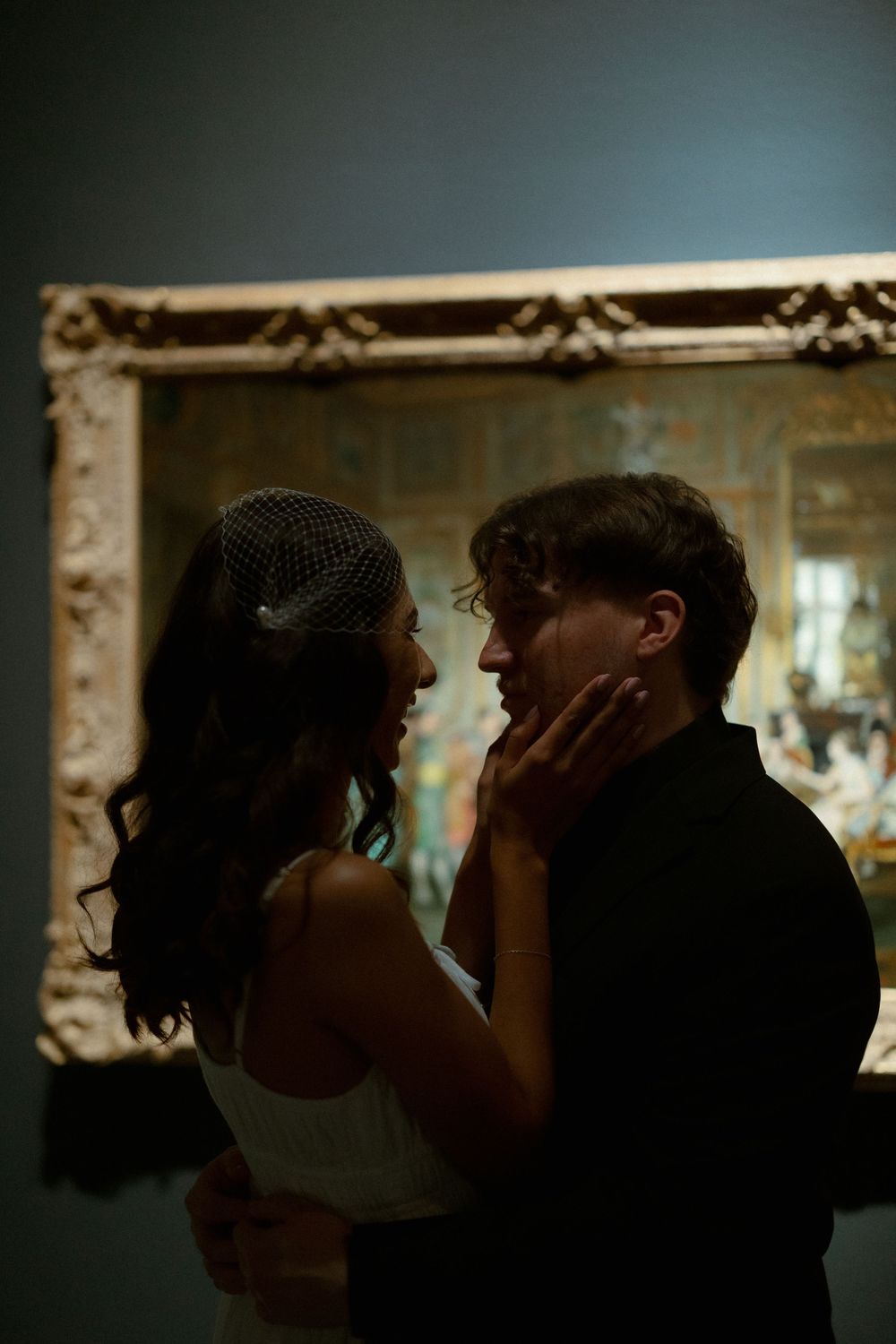 A romantic couple shares an intimate moment in front of an ornate gold frame in a dimly lit room.